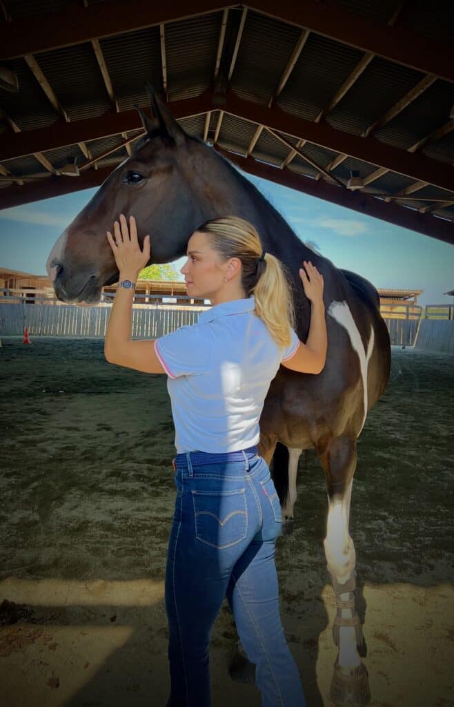 Câlin entre cavalière blonde et cheval bai en écurie Joueuse blonde en polo blanc et jean caresse la tête et le flanc d'un cheval bai avec une marque blanche dans une écurie.